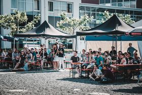 Blick auf den Schotterplatz des HTWG-Campus. Auf dem Schotter stehen zwei schwarze Pavillonzelte, unter deren Dächer Menschen an Biertischgarnituren sitzen.
