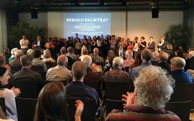 Präsentation in einem Raum voller Menschen von der letzten Sitzreihe aus fotografiert. Auf der aktuellen Präsentationsfolie steht "Rebuild Palmyra".