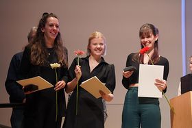 Das Bild zeigt die drei Preisträgerinnen mit ihren Urkunden und Blumen in der Hand auf der Bühne während der Preisverleihung