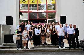 Große Gruppe von etwa 25 Personen steht auf den Außentreppen eines Gebäudes, mehrere junge Preisträgerinnen in der vorderen Reihe halten Auszeichnungen in der Hand. Im Hintergrund ist der Gebäudeeingang mit großen Fensterfronten zu sehen.