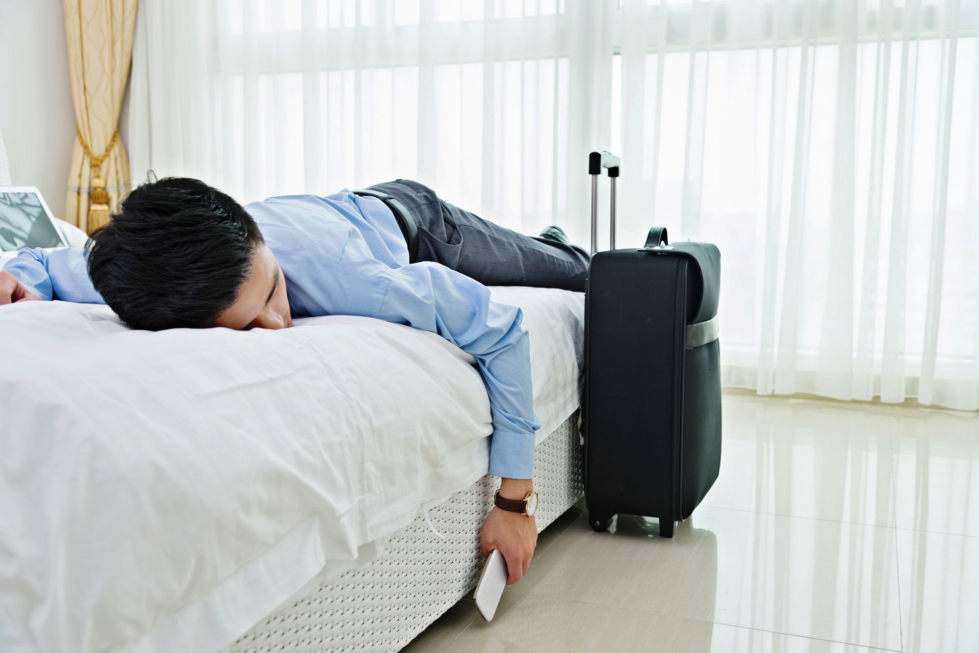 A young Asian manager in a blue shirt and business pants is lying exhausted on a bed in a hotel room. On the floor next to him is his suitcase. His arm is hanging off the bed towards the floor, his smartphone in his hand. 