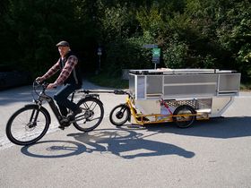 Ein Mann fährt auf einem Ebike, das einen Anhänger zieht. Auf dem Anhänger ist eine Aluminium-Box befestigt.