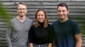 Das Bild zeigt zwei junge Männer und eine junge Frau in der Mitte. Sie stehen vor einer Holzwand und lächeln freundlich in die Kamera. 