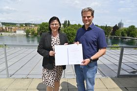 Ein Mann und eine Frau stehen auf einer Dachterrasse und halten lächelnd eine Urkunde hoch