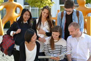 Eine Gruppe Studierender sitzt vor den gelben Figuren auf dem HTWG Campus und schaut in die Unterlagen einer Studierenden.