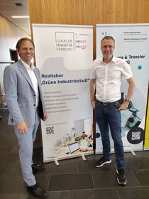 Das Bild zeigt Gunnar Schubert und Stefan Stieglat vor einem Aufsteller zum Reallabor Grüne Industriestadt