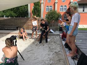 Ein Videodreh in der Strandbar vor der HTWG: Drei Studierende sind vor der Kamera. Eine davon sitzt im Liegestuhl. Ein weiterer steht mit einem Strohhut und einer Badehose bekleidet im Sand und hat ein Plakat vor der Brust. Der dritte spielt auf einer Gitarre. Vor ihnen im Sand sitzt eine Studentin hinter einer Kamera. Weitere stehen am Rand des Geschehens.