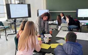 Ein Foto aus einem Vorlesungsraum, in dem eine Frau und mehrere Schüler*innen an Gruppentischen zusammenarbeiten, während im Hintergrund Präsentationsbildschirme vor einer grünen Tafel zu sehen sind.
