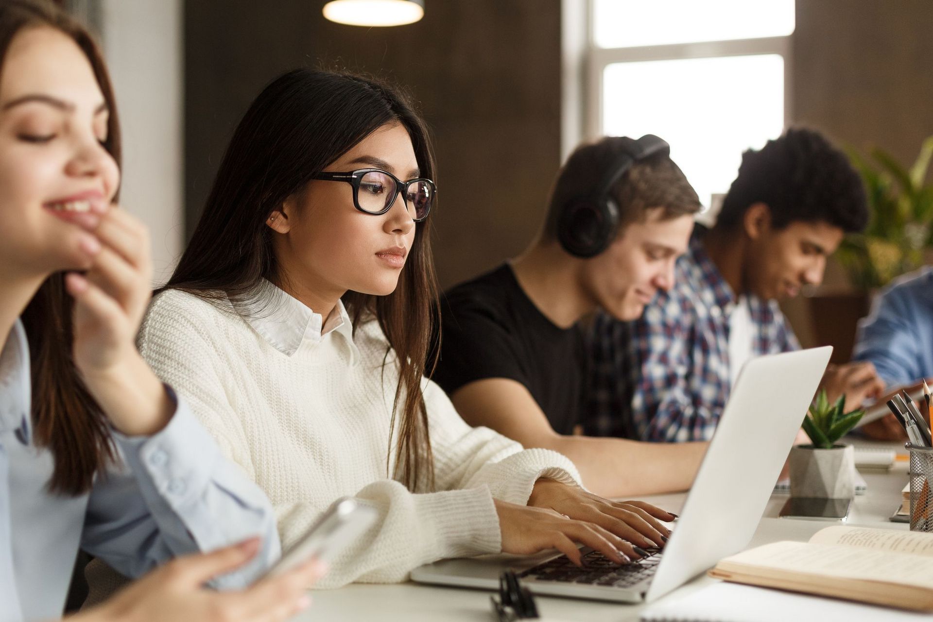 Gruppe von Studierenden (unterschiedliche Nationalitäten), die nebeneinander in der Vorlesung sitzen. Eine Studentin prüft ihr Mobiltelefon, eine andere schaut in ihr Notebook, ein weiterer Student hat Kopfhörer auf.