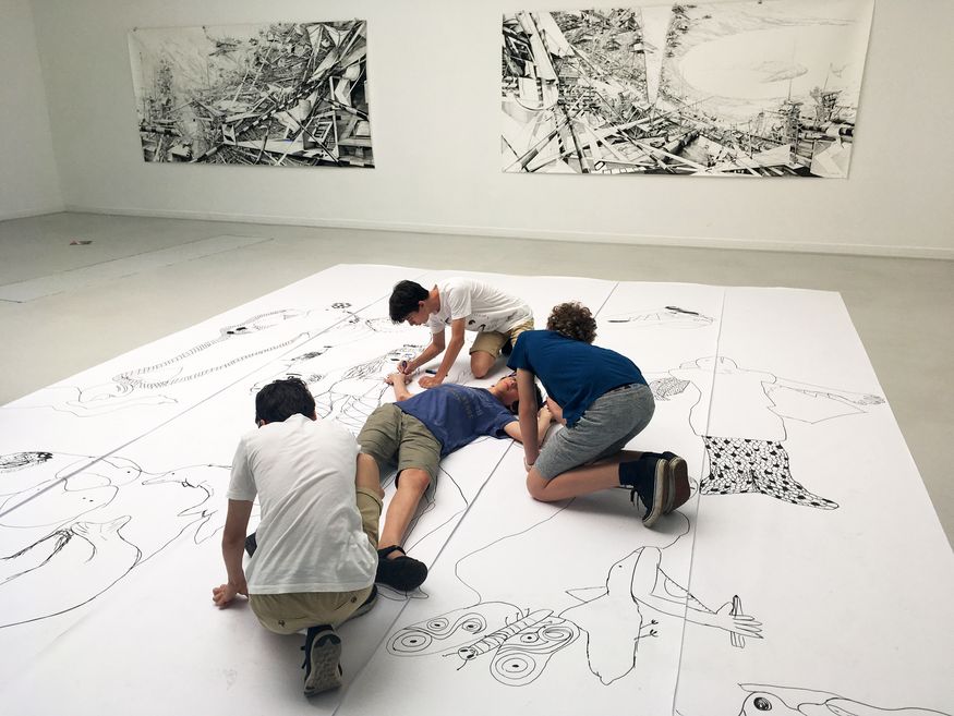 Ein Schüler liegt auf dem Rücken auf einer großen Papierfläche. Drei Schüler knien um ihn herum und zeichnen seine Konturen auf das Papier ab.
