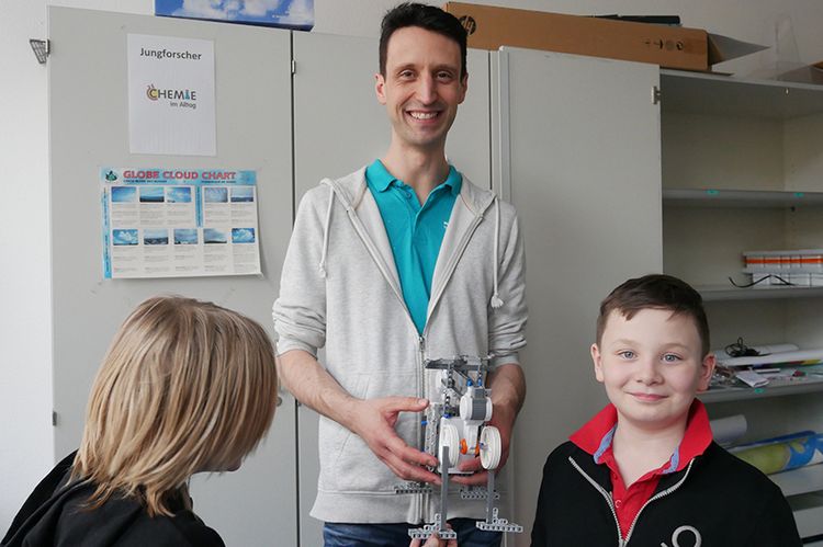 Student Sebastian Rewig steht vor einem großen, weißen Schrank, hält einen Mind-Storm-Roboter in den Händen und blickt lächelnd in Richtung Kamera. Vor ihm stehen zwei Schüler, von einem sieht man lediglich den Hinterkopf, der zweite blickt ebenfalls lächelnd in die Kamera.