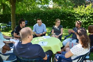 Sieben Personen sitzen in grünem Garten an einem Tisch und unterhalten sich