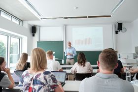 Das Bild zeigt einen hellen Seminarraum. Studierende sitzen mit dem Rücken zur Kamera und schauen nach vorne zu ihrem Professor. Hinter ihm ist ein Diagramm auf die Wand projiziert.