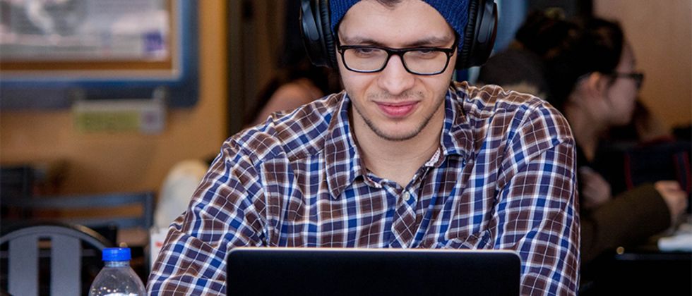 Ein junger Mann mit Mütze und großen Kopfhörern arbeitet im Café an seinem Laptop.