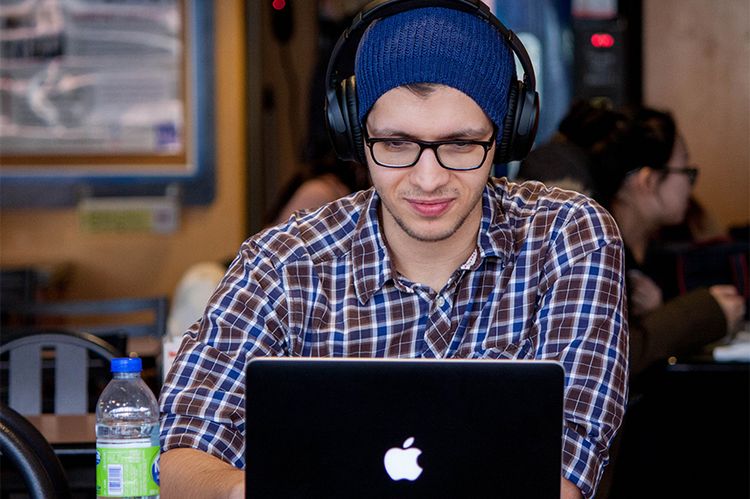 Ein junger Mann mit Mütze und großen Kopfhörern arbeitet im Café an seinem Laptop.