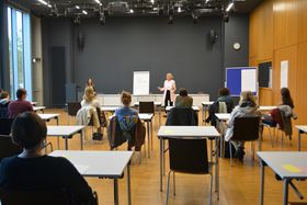Blick von hinten in einen großen Saal. Mit großem Abstand sitzen Frauen an Einzeltischen. Vorne spricht Gleichstellungsbeauftragte Prof. Dr. Kerstin Schaper-Lang mit Blick ins Publikum.