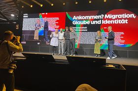 Bühne bei einer Preisverleihung: Sechs Personen stehen vor einer großen LED-Wand, auf der „Ankommen? Migration, Glaube und Identität" und „Semesterarbeit HTWG Konstanz" mit der Auszeichnung Gold angezeigt werden. Im Vordergrund fotografiert ein Fotograf die Szene.