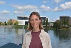 Eine junge Frau mit blonden Haaren lächelt in die Kamera. Im Hintergrund ist der Seerhein und ein weißes Gebäude (Bodenseeforum) zu sehen