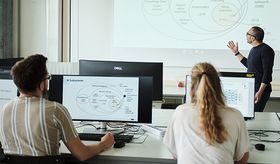 Ein Mann steht vor einer Präsentation, zwei Studierenden hören zu und schauen zu ihm