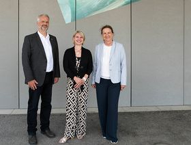 Ein Mann und zwei Frauen stehen vor einer grauen Betonwand. Sie sind schick gekleidet.