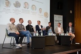 Sieben Männer auf einer Bühne bei einer Paneldiskussion, im Hintergrund eine Leinwand mit einer Präsentation