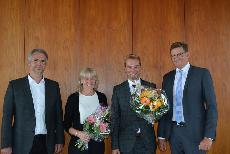 Manfred Schnell, Prof. Dr. Beate Bergé, Prof. Dr. Oliver Haase und Prof. Dr. Carsten Manz posieren für ein Gruppenfoto vor einer Holzwand. Beate Bergé und Oliver Haase halten Blumensträuße in den Händen.  