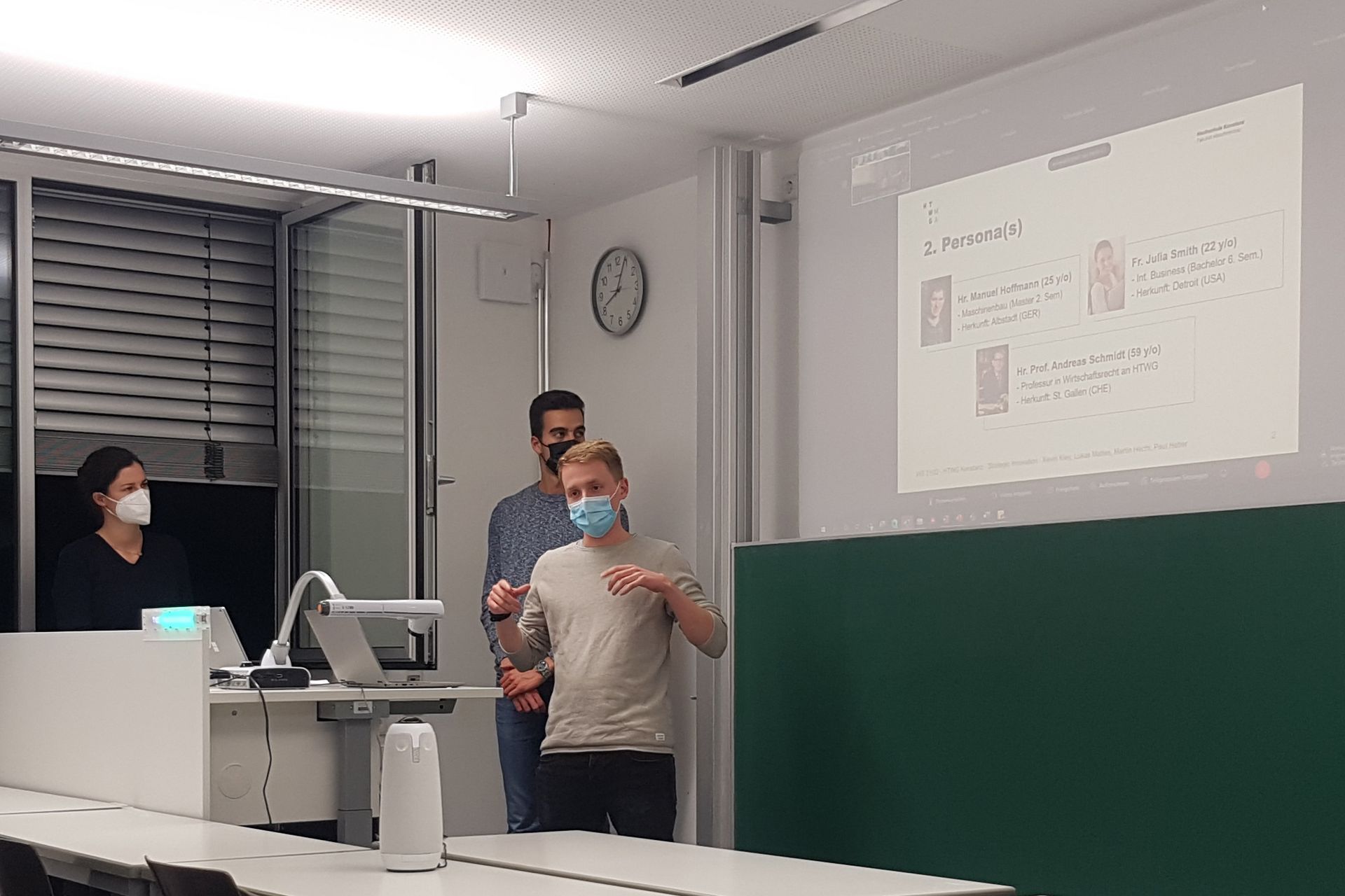 Zwei Studenten stellen vor einer Projektion ihrer Präsentation ihr Projekt vor. Auf einem Tisch vor ihnen, steht eine 360-Grad-Kamera.