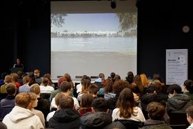 Rund 90 Schülerinnen und Schüler sitzen in einem Vortragssaal. Sie sind von hinten zu sehen. Vorne am Rednerpult steht Studienberaterin Sandra Flügel. 