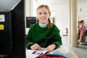 Das Bild zeigt eine Schülerin des Heinrich-Suso-Gymnasiums an einem Computerarbeitsplatz. Sie trägt einen grünen Pullover, der die Aufschrift des Gymansiums aufgedruckt hat. Ihre Haare hat sie zu zwei blonden Zöpfen gebunden.