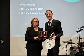 Eine Frau und ein Mann blicken lächelnd in die Kamera, sie halten ein Newton-Pendel und einen überdimensionierten Schlüssle mit "HTWG"-Anhänger in der Hand
