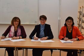 Katharina Holzinger, Monika Knill und Sabine Rein sitzen an einem Holztisch und unterzeichnen jeweils einen Vertrag. Sie lächeln dabei in die Kamera.