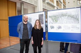 Ein junger Mann und eine junge Frau posieren vor Infotafeln mit Stadtplänen fürs Foto.