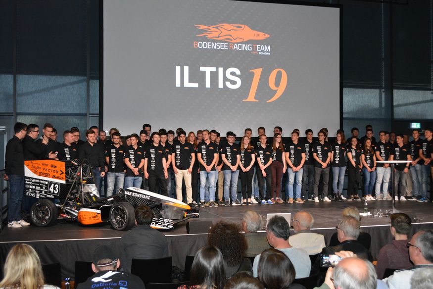 Eine Gruppe von zirka 50 jungen Menschen in schwarzen T-Shirts steht vor einem Bildschirm auf einer Leinwand und blickt ins Publikum. Auf der Leinwand steht in weißen Buchstaben Iltis 19.