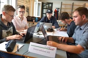 Fünf junge Männer sitzen um einen Tisch. Darauf stehen Laptops und ein Schild mit der Aufschrift "priME CUP - Unternehmen A2".