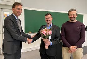 Foto zeigt drei Personen, im Hintergrund ist eine grüne Tafel. Die Person links reicht der Person in der Mitte die Hand. Die Person in der Mitte hält einen Blumenstrauß in der Hand. Die Person rechts steht daneben.