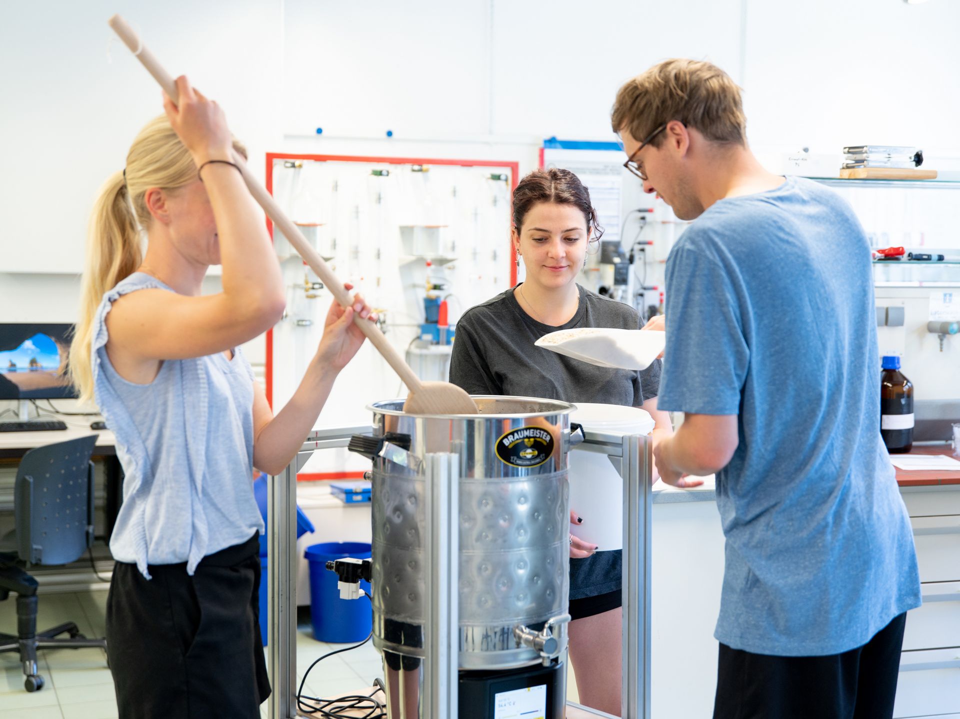 Drei Student*innen arbeiten im Labor mit einem kleinen Braukessel.