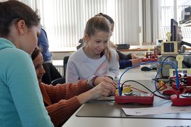 In einem Seminarraum sitzen Mädchen an einem Tisch. Sie arbeiten zusammen mit roten Kabelnd und Buchsen.