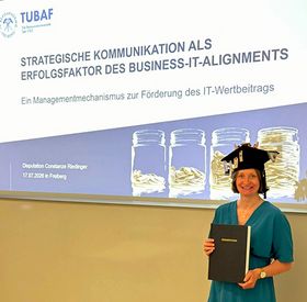 Person mit Doktorhut und gebundener Dissertation vor Präsentationsfolie mit Titel (abgekürzt) „Strategische Kommunikation als Erfolgsfaktor des Business-IT-Aligments“