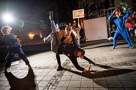 Ein junger Mann und eine junge Frau beim wilden Tanz auf der Straße. Um sie herum tanzen wietere Personen teils mit Protestplakaten, teils mit Bauhelmen auf dem Kopf.