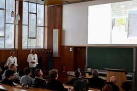 Zwei Männer stehen in einer Aula vor Publikum und schauen auf eine Präsentation
