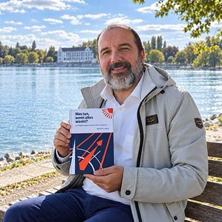 Prof. Dr. Erdal Yalcin präsentiert seine Buchneuerscheinung auf einer Bank am Ufer des Bodensees; im Hintergrund die Silhouette der Stadt Konstanz. Foto: Luise Yalcin.