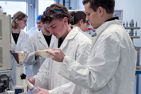 Studierende in weißen Laborkitteln im Labor. Ein Student hält eine Schaufel mit Haferkörnern in Richtung eines Trichters.