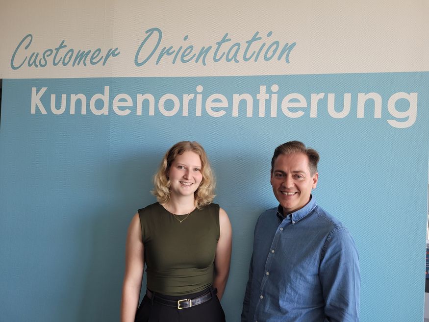 In front of a wall with "Customer Orientation / Kundenorientierung" in white and blue on the wall, stands Dana Karletshofer with the head of CSP Market Management Reinhold Unmuth.