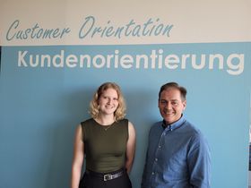 In front of a wall with "Customer Orientation / Kundenorientierung" in white and blue on the wall, stands Dana Karletshofer with the head of CSP Market Management Reinhold Unmuth. 