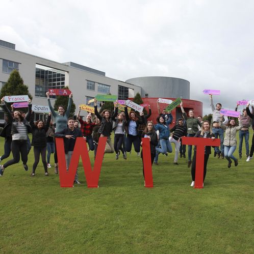 Logo des Waterford Insitutes of Technology im Vordergrund mit Austauschstudierenden, die die Namen ihrer Herkunftsländer auf Schildern in die Höhe halten. Im Hintergrund Gebäudeansicht des WTS