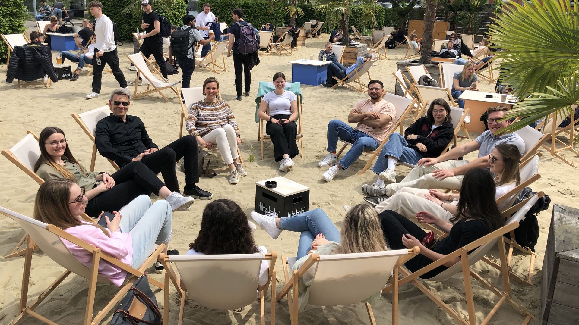 Eine Gruppe von Studierenden und zwei Professor*innen bildet einen Stuhlkreis aus Liegestühlen. Sie sitzen auf Sand, in der Strandbar der HTWG Konstanz.