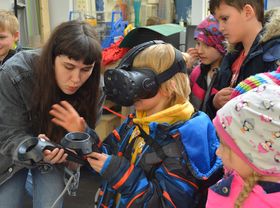 Ein Kind umringt von anderen Kindern: Es trägt eine VR-Brille und hält ein Steuerungsgerät in der Hand. Eine junge Frau beugt sich zu ihm hinab.