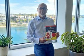 Ein Mann mit einem Buch mit dem Titel "Juristische Schemata für Dummies" lächelt in die Kamera. Im Hintergrund sieht man einen Fluss und Häuser.