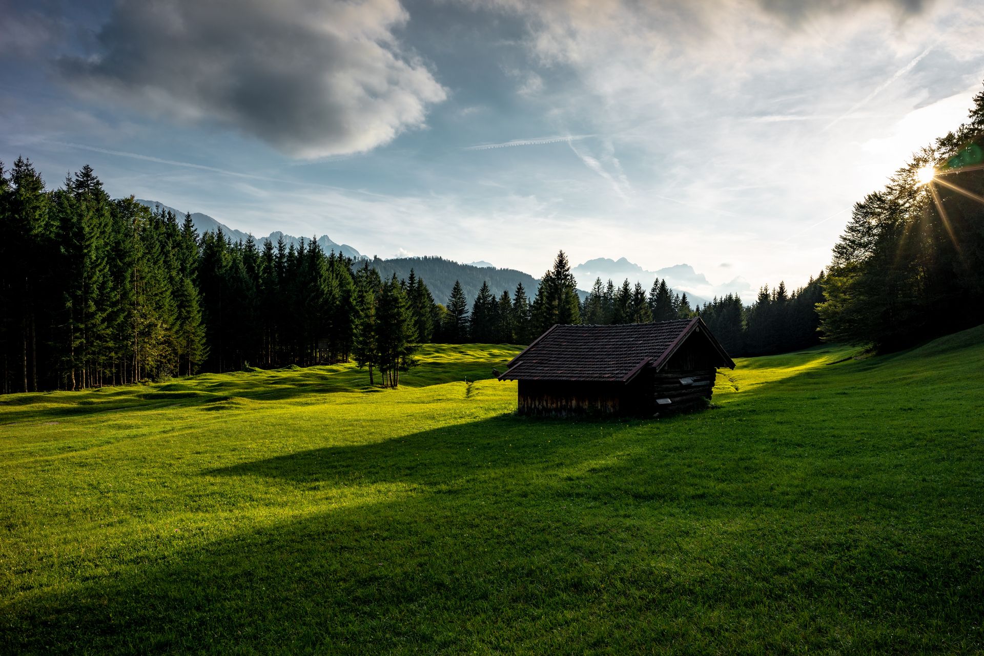 Eine Holzhütte steht auf einer grünen Wiese. Im Hintergrund sind Wald und die Alpen zu sehen.