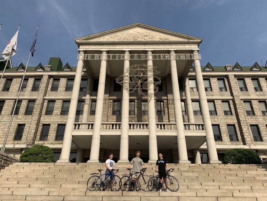3 junge Männer stehen mit ihren Fahrrädern auf einer Treppe vor einem großen Gebäude mit Säuleneingang